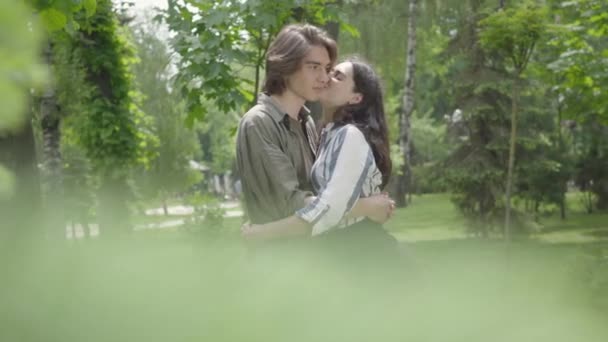 Nettes junges Paar, das sich im Hintergrund zärtlich umarmt und küsst, unscharfe Blätter im Vordergrund. glückliche Mädchen und Jungen, die Zeit miteinander im Park verbringen. Freizeit im Freien — Stockvideo