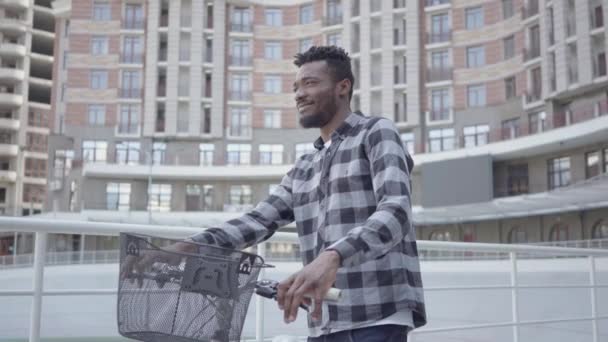 都市建築を背景に自転車で立つハンサムなアフリカ系アメリカ人男性の肖像画。男は屋外で良い天気で休んでいる — ストック動画