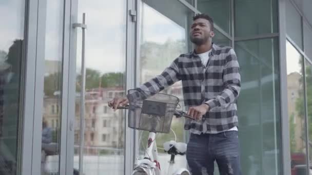 Красивий впевнений афроамериканець людини в повсякденний одяг, що ведуть свій велосипед біля магазину вікна в місті. Хлопець витрачає час на свіжому повітрі, активний спосіб життя — стокове відео