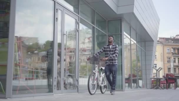 Retrato seguro atractivo hombre afroamericano de pie con bicicleta sobre el fondo de la arquitectura urbana. El tipo está descansando con buen tiempo al aire libre. — Vídeo de stock