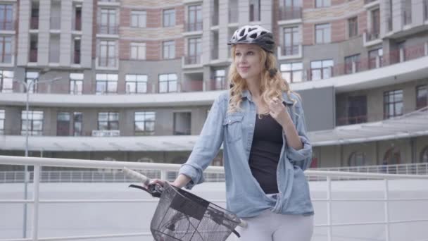 Portret schattig blond vrouw in fietshelm staande met fiets tegen de achtergrond van stedelijke architectuur. De dame is op fietspad in de stad. Actieve levensstijl. — Stockvideo