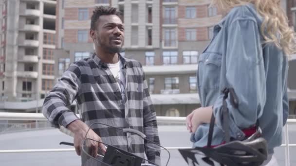 Portret knappe Afro-Amerikaanse man staand met fiets tegen de achtergrond van stedelijke architectuur praten met een schattige aantrekkelijke blonde vrouw. Paar vrienden chatten buitenshuis — Stockvideo