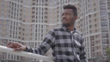 Şehir mimarisinin arka planına karşı duran yakışıklı Afro-Amerikan bir adamın portresi. Adam iyi havalarda açık havada dinleniyor, arka planda gökdelen