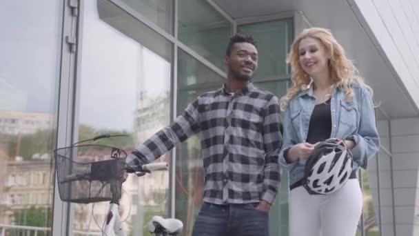 Knap zelfverzekerd Afro-Amerikaanse man in Casual kleding leidt zijn fiets in de buurt van de etalages in stad praten met blanke vriendin. Het internationale echtpaar besteedt tijd samen buiten — Stockvideo