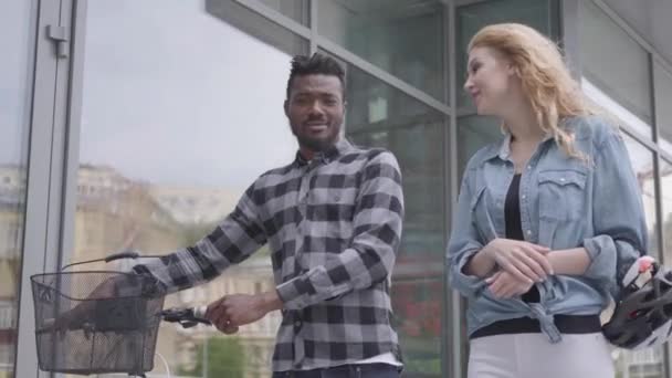 Portret knap zelfverzekerd Afro-Amerikaanse man in Casual kleding leidt zijn fiets in de buurt van de etalages in stad praten met blanke vriendin. Het internationale koppel besteedt tijd samen — Stockvideo