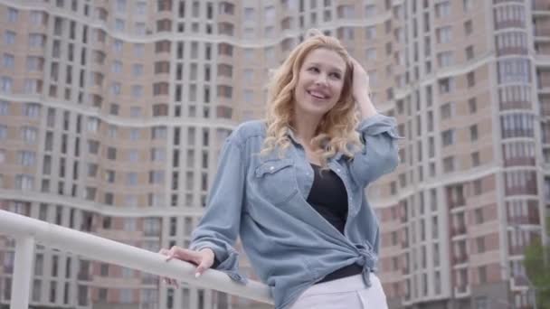 Ganska säker blond kvinna i Jeansjacka poserar framför skyskrapa, ser bort leende. Stads livsstil, stadsliv. Attraktiv stadsdweller med hög byggnad i bakgrunden — Stockvideo