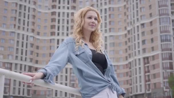 高層ビルの前でポーズをとるジーンズジャケットを着たかなり自信に満ちたブロンドの女性を肖像画し、笑顔で見つめている。都会のライフスタイル、都会生活。背景に高い建物を持つ魅力的な都市の人々 — ストック動画