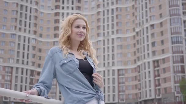 Portrait mignon confiant femme blonde en jean veste posant devant le gratte-ciel, regardant loin en souriant. Mode de vie urbain, vie urbaine. Attrayant citadin avec bâtiment haut sur le fond — Video