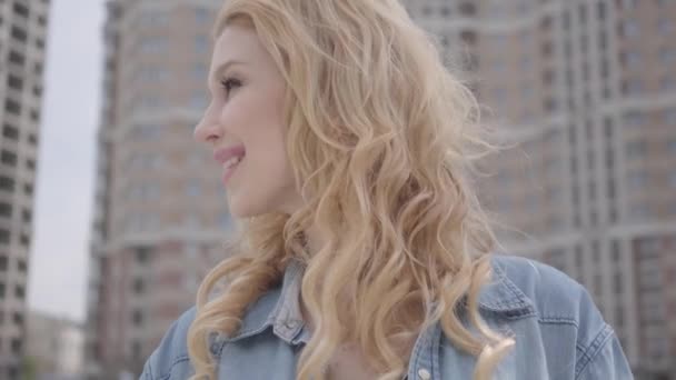 Portrait d'une jolie femme blonde confiante et souriante parlant avec un interlocuteur méconnaissable devant un gratte-ciel. Style de vie urbain. Femme citadine avec bâtiment haut sur fond — Video
