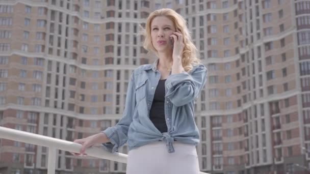 超高層ビルの前で携帯電話で話すかわいい笑顔の自信ブロンド女性の肖像画。都会的なライフスタイル。背景に高い建物を持つ女性都市の人々 — ストック動画
