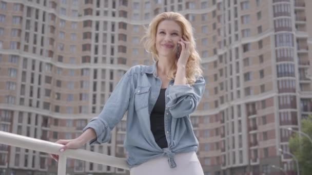 Fiduciosa donna bionda sorridente in giacca di jeans che parla al cellulare di fronte al grattacielo. Stile di vita urbano. Abitante di città femminile con alto edificio su sfondo — Video Stock