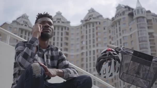 Hombre afroamericano guapo sentado cerca de su bicicleta en el fondo de la arquitectura urbana. El tipo hablando por celular al aire libre, rascacielos en el fondo. Ocio de verano — Vídeos de Stock