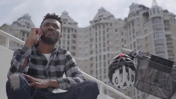 Retrato bonito afro-americano sentado perto de sua bicicleta no fundo da arquitetura urbana. O tipo a falar ao telemóvel ao ar livre, um arranha-céu no fundo. Lazer de verão — Vídeo de Stock