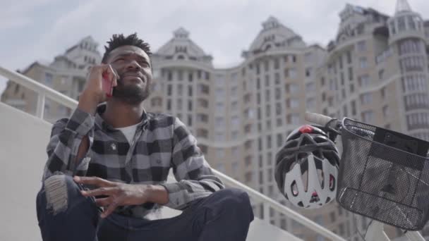 Ritratto carino bell'uomo afroamericano seduto vicino alla bicicletta sullo sfondo dell'architettura urbana. Il tizio che parla al cellulare all'aperto, il grattacielo sullo sfondo. Tempo libero estivo — Video Stock