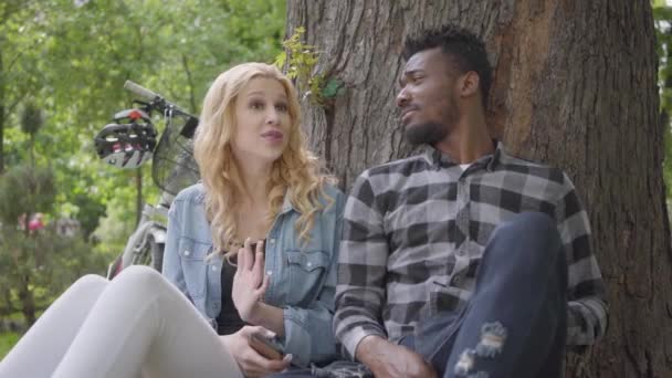 Portret confidient mooie blonde vrouw en knappe Afro-Amerikaanse man praten zittend onder een oude boom in het Park. De moderne fiets staat in de buurt. Het internationale paartijd doorbrengen — Stockvideo