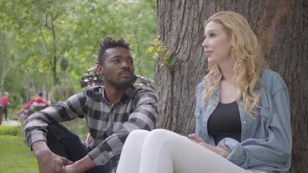 Retrato confiado mujer rubia bonita y apuesto hombre afroamericano hablando sentado debajo de un viejo árbol en el parque. La bicicleta moderna de pie cerca. La pareja internacional pasa tiempo — Vídeos de Stock