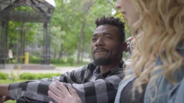 Portre sırdaş güzel sarışın kadın ve parkta eski bir ağacın altında oturan yakışıklı Afrikalı Amerikalı adam konuşuyor. Modern bisiklet yanında duruyor. Uluslararası çift zaman harcama