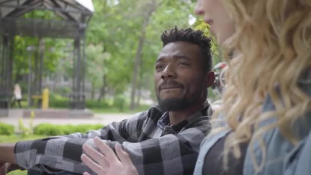 Portret confidient całkiem blond kobieta i przystojny African American mężczyzna mówi siedzi pod starym drzewem w parku. Nowoczesny rower stojący w pobliżu. Międzynarodowa para spędza czas — Wideo stockowe