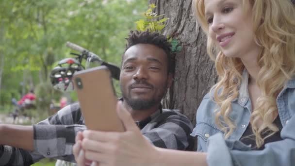 Szép szőke nő és szép afro-amerikai férfi ül egy régi fa a parkban. A hölgy bemutató képek barátja a mobilját a nemzetközi pár időtöltést a szabadban — Stock videók
