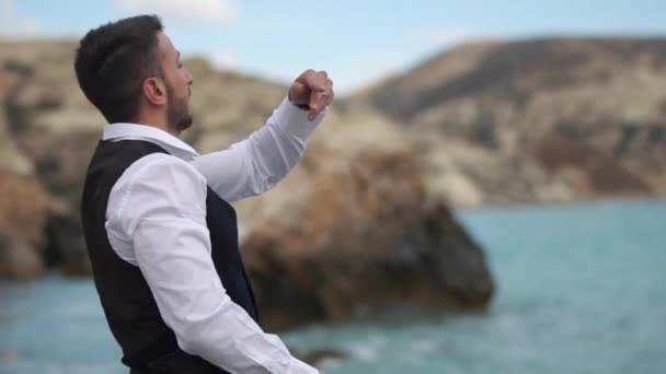 Barbudo joven guapo cerca del mar y las montañas arrojando piedras en el agua. En cámara lenta. Chipre. Paphos. — Vídeos de Stock