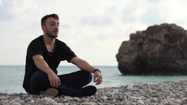 Gözleri kapalı genç bir adam, deniz manzarasının yanında yoga tarzında oturuyor ve rahatlıyor. Bir kayanın ve denizin arka planında meditasyon yapan yakışıklı bir adam. Zaman turları. Kıbrıs. Papolar