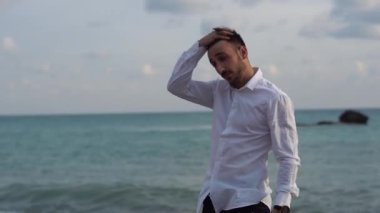 Genç şık adam sahilin kenarında duruyor ve koyu renk saçlarına eliyle dokunuyor. Yakışıklı bir adam eliyle saçını düzeltiyor. Kıbrıs. Papolar