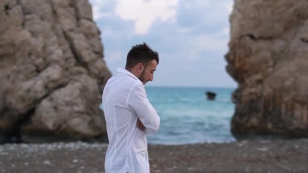 年轻英俊的男子在岩石之间的海滩上走在多风的海滨附近。 塞浦路斯。 帕福斯 — 图库视频影像