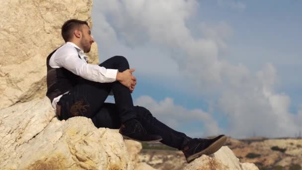 Hombre elegante guapo sentarse en las rocas y con ganas de una vista increíble. Chipre. Paphos. — Vídeos de Stock