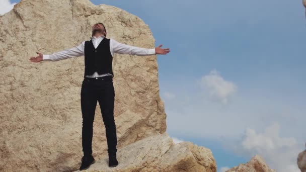 Joven hombre con estilo que le dice a la gente lo feliz que está de estar en este hermoso lugar con una vista increíble. Un tipo libre de éxito encima de una gran piedra. Chipre. Paphos. — Vídeo de stock