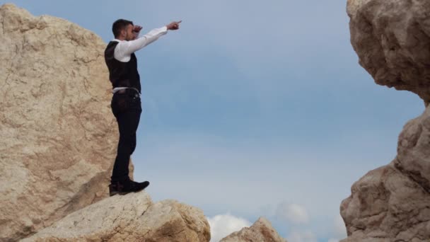 Bello uomo elegante in stoffa fantasia in piedi sulle rocce e waching una vista incredibile, mostrando qualcosa alla gente con il dito. Cipro. Paphos — Video Stock