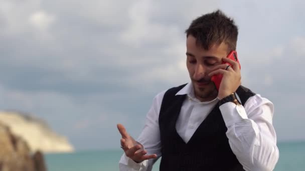 Retrato de un hombre molesto y frustrado hablando en un teléfono móvil cerca del mar con una vista impresionante. Un hombre de negocios hablando por el celular al aire libre. Chipre. Paphos. — Vídeos de Stock
