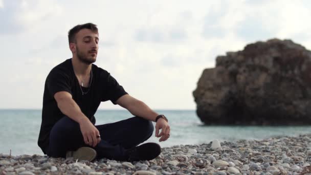 Joven con los ojos cerrados sentado y relajante en estilo yoga cerca de increíble vista al mar. Un chico guapo meditando sobre el fondo de una roca y el mar. El tiempo pasa. Chipre. Paphos. — Vídeo de stock