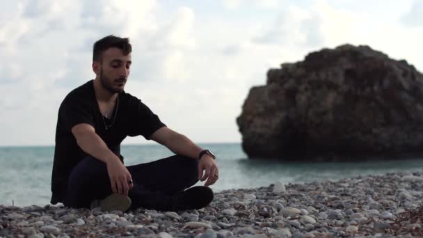 Bonito homem meditando no fundo de uma rocha e do mar. O tempo passa. Jovem com os olhos fechados sentado e relaxante em estilo ioga perto de vista mar incrível. Chipre. Paphos — Vídeo de Stock