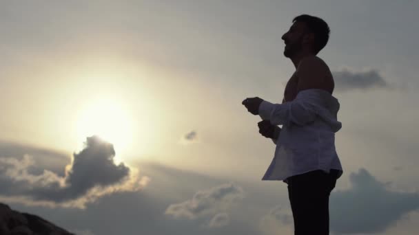 Un joven al atardecer se quita su camisa clásica blanca y disfruta felizmente de una vista y puesta de sol increíbles. Turista feliz en la cima de la roca, victoria y propósito . — Vídeo de stock