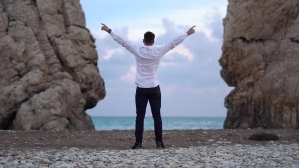 El hombre con una camisa blanca y jeans oscuros tirando sus manos al costado, disfrutando de la libertad, cree en el éxito futuro. Turista feliz en la cima de la roca, victoria y propósito. Chipre. Paphos. — Vídeos de Stock