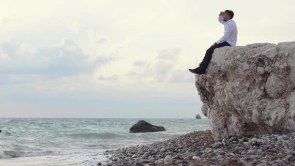 Hombre guapo y elegante sentado en las rocas y con una vista increíble mientras muestra algo a la gente con su dedo. Chipre. Paphos. — Vídeos de Stock