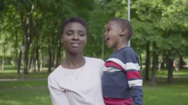 Yeşil parkta oğlunu kollarında tutan Afro-Amerikan kadının portresi yakın. Sevimli çocuk annesi ile açık havada vakit geçiriyor. Sevgi dolu aile, kaygısız çocukluk