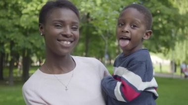Sevimli Afro-Amerikan kadın yeşil parkta kollarında yakın onu komik oğlu tutarak portresi. Sevimli çocuk kameraya dilini gösteriyor, annesi gülüyor. Sevgi dolu aile, kaygısız