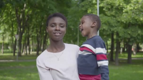 Retrato de una mujer afroamericana sosteniendo a su hijo en brazos en el parque verde de cerca. Lindo niño pasando tiempo al aire libre con su madre. Familia cariñosa, infancia despreocupada — Vídeos de Stock