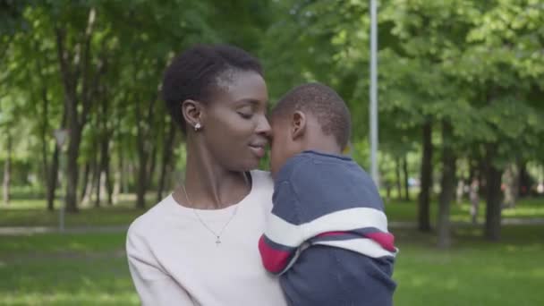 Retrato mulher afro-americana bonita segurando seu filho em seus braços no parque verde de perto. Criança bonito passar tempo ao ar livre com sua mãe. Família amorosa, infância despreocupada — Vídeo de Stock