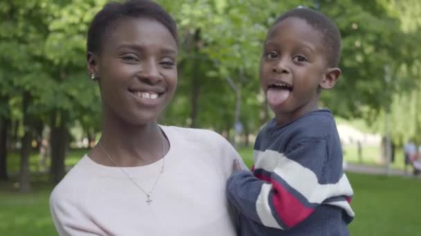 Πορτρέτο του αξιολάτρευτο αφρικανική Αμερικανίδα γυναίκα κρατώντας το αστείο γιο της στα χέρια της στο πράσινο πάρκο κοντά. Χαριτωμένο παιδί που δείχνει τη γλώσσα του στην κάμερα, η μητέρα γελάει. Στοργική οικογένεια, ανέμελη — Αρχείο Βίντεο