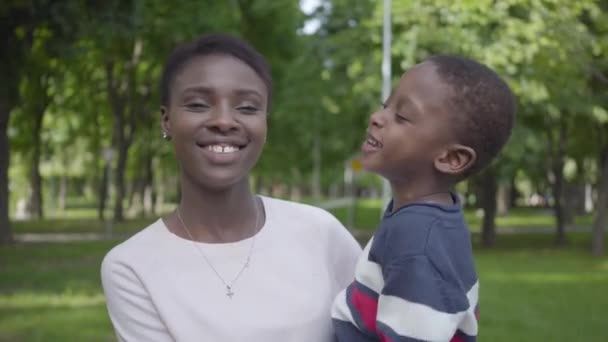 Retrato linda mujer afroamericana sosteniendo a su hijo divertido en sus brazos en el parque verde de cerca. Familia cariñosa, infancia despreocupada — Vídeos de Stock