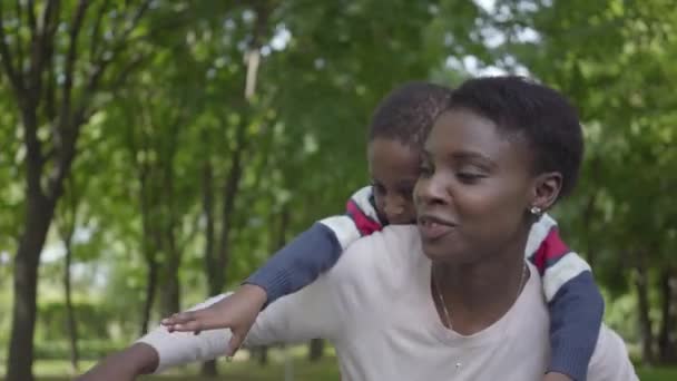 Портрет афроамериканской женщины, держащей сына на спине в зеленом парке с раскинутыми в сторону руками. Милый ребенок проводит время с матерью на улице. Любящая семья, беззаботное детство — стоковое видео