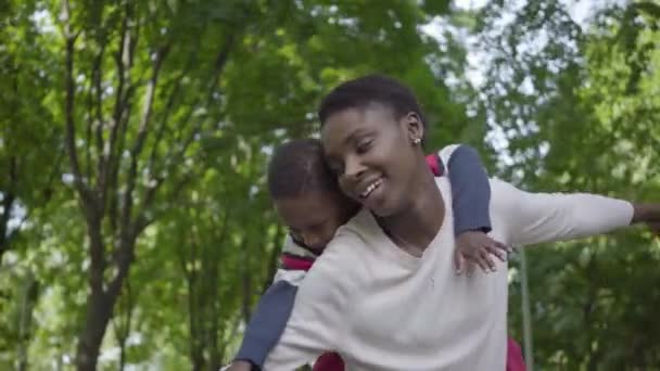 녹색 공원에서 아들을 안고 있는 귀여운 아프리카 계 미국인 여성의 초상화가 팔을 뻗어 옆으로 뻗어 있습니다. 귀여운 아이는 야외에서 어머니와 함께 시간을 보내고 있습니다. 사랑하는 가족, 평온한 — 비디오