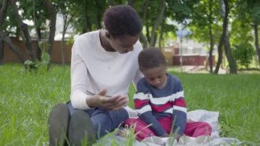 Portre çekici Afrikalı-Amerikalı kadın parkta bir oyuncak ile oynarken küçük oğlu ile battaniye üzerinde oturan. Genç anne dışarıda çocuğuyla vakit geçiriyor. Sevgi dolu aile üzerinde