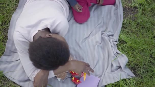 Hermosa mujer afroamericana con el pelo corto acostado en la hierba en el parque con su hijo jugando con el juguete transformador de plástico. La madre con un hijo afuera. Amando a la familia en el picnic. Vista superior — Vídeo de stock