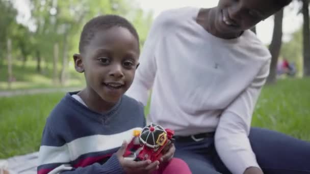 Portre güzel Afro-Amerikan güzel genç kadın parkta bir oyuncak ile oynarken küçük oğlu ile battaniye üzerinde oturan. Genç anne dışarıda çocuğuyla vakit geçiriyor. Sevgi dolu aile üzerinde — Stok video
