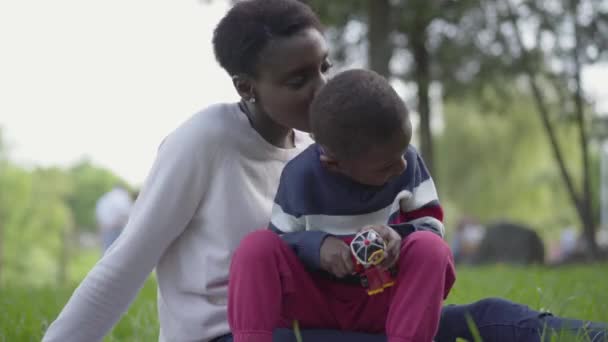 Little cute African American boy playing with plastic toy in the park, sitting on mothers laps close up. The woman and her child resting outdoors. Loving family on the picnic — Stock Video