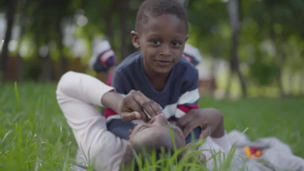 Pretty African American woman lying on the grass in the green park, her little son lying on top of her. Mother and boy playing outdoors close-up. Motherhood concept — Stock Video