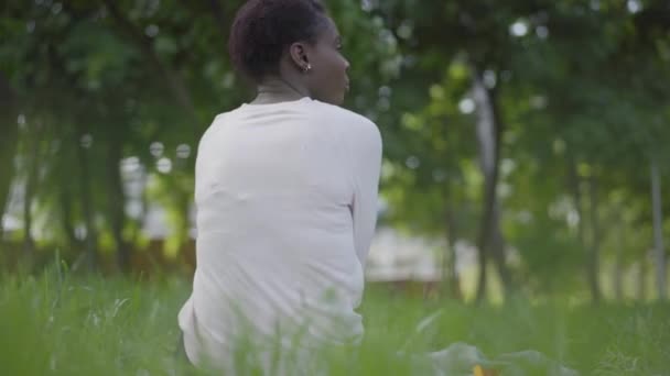 Vista trasera de la mujer afroamericana sentada en el césped observando a su pequeño hijo jugando y corriendo alrededor de los árboles. Madre y niño pasando tiempo al aire libre — Vídeo de stock